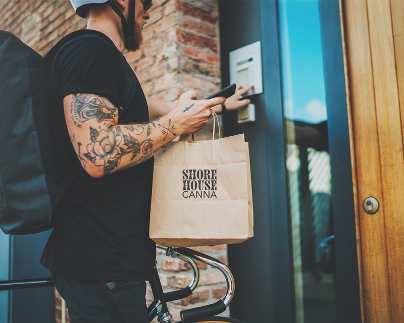 Shore House Canna West Cape May dispensary delivery: a tattooed courier in a helmet stands by a bicycle, holding a Shore House Canna paper bag and pressing an intercom at a residence